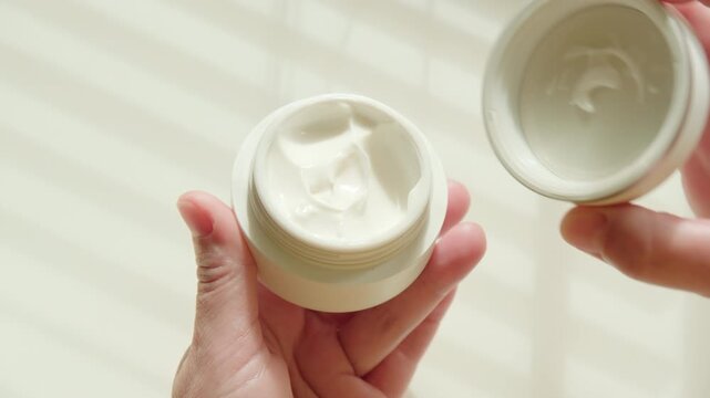 Woman hand opening lid of jar with white cosmetic cream on background of dressing table with shadow from blinds on window from sunlight in morning closeup