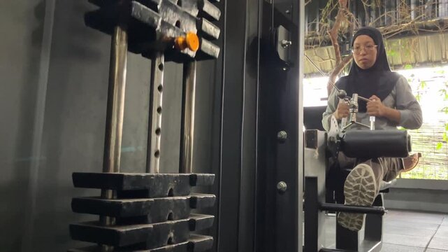 Slow motion video of a hijab-wearing woman performing a strength training exercise on a gym cable machine, focusing on the moving weight stack.