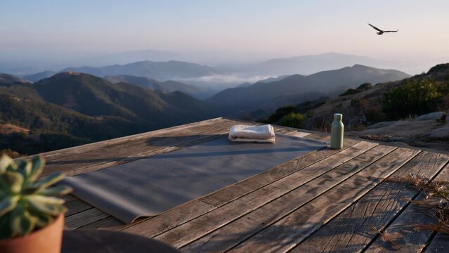 A yoga mat with a towel and water bottle on a wooden deck overlooking mountains with platform against backdrop backdrop mountain ranges folded towel