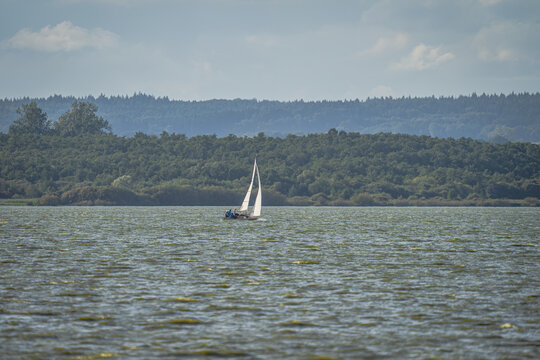 Ein kleines Segelboot mit wei&szlig;em Segel gleitet &uuml;ber das leicht gekr&auml;uselte, gr&uuml;nlich schimmernde Wasser des D&uuml;mmer-Sees. Im Hintergrund erstreckt sich ein dichter, dunkler Waldstreifen unter einem hel