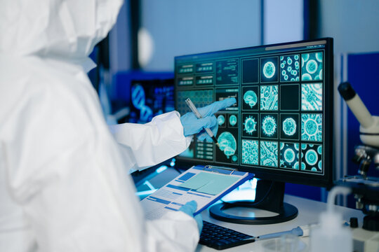 Medical researcher in protective suit reviewing and recording data from multi screen digital displays inside research laboratory.