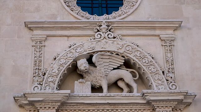 Stone winged lion relief stands above the Chiesa di San Marco doorway, framed by ornate baroque carvings in soft daylight.