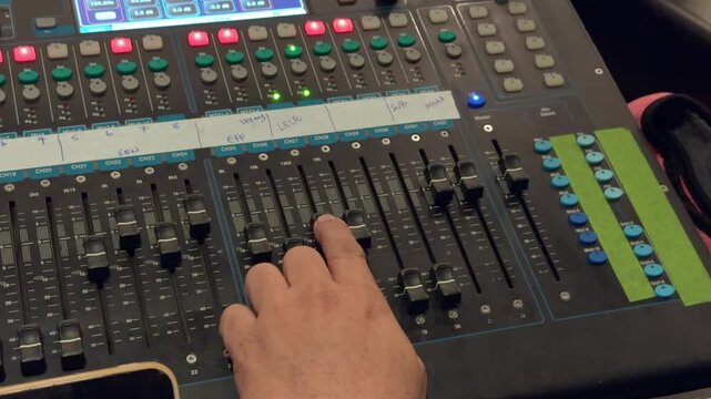Close view from above of a hand adjusting channel faders on a digital sound mixing desk with rows of rotary knobs illuminated controls and handwritten labels