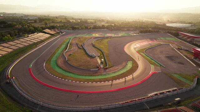 Drone pans across cyclists riding around a motorsport circuit under warm sunrise light.