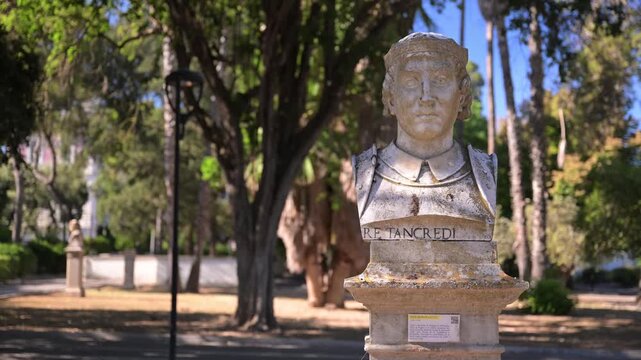 Marble bust of King Tancred stands on a pedestal in Villa Comunale Giuseppe Garibaldi, framed by sunlit trees and paths in a quiet city park.