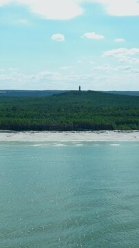 A vertical aerial shot showing the Baltic Sea coast with a white sand beach, lush green pine forest, and the historic Stilo lighthouse on a hill in Poland.