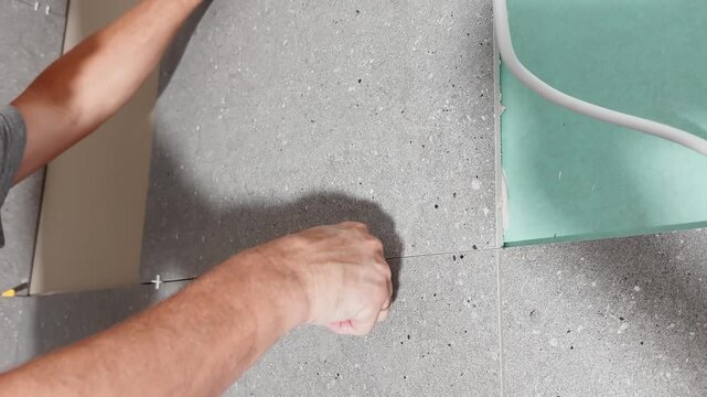 Worker positions a large gray wall tile on a prepared interior surface, aligning it by hand during renovation and professional ceramic wall finishing.