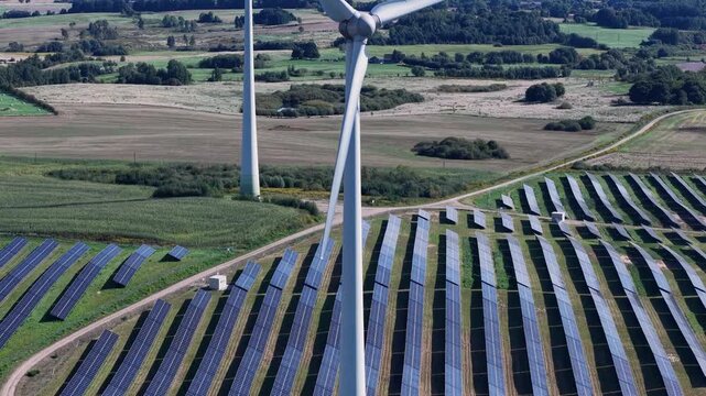 Aerial footage captures rows of solar panels and towering wind turbines operating together on lush farmland, highlighting clean, sustainable energy in a rural landscape under clear daylight.