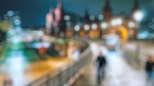 A blurred nighttime cityscape featuring lights and architecture along a riverbank, creating an atmospheric and moody urban scene.