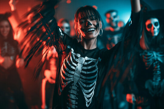 Skeleton at Halloween Costume Party Smiling Woman Dancing with Raised Arms and Skull Makeup Among Costumed Friends