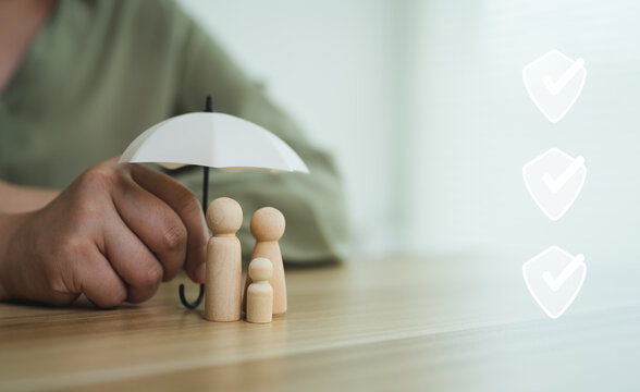 Concept of family insurance protection with wooden doll figures under small umbrella and safety checkmark icon.