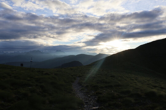 Mountain hiking trail at Passo delle Sette Crocette with sunbeams breaking through dramatic clouds at sunrise, Prealpi Bresciane, Italy