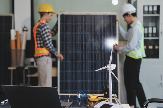 Two Asian engineers wearing hardhats and safety vests discussing solar panel installation and renewable energy project in the office.