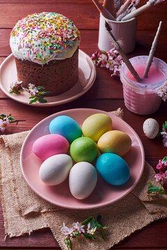 Selection of colourful painted easter eggs on apink plate. There is also an Easter cake, and paints & paint brushes on the wooden table.