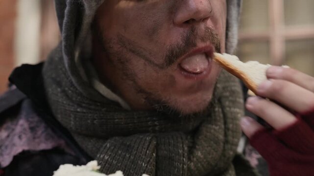 Caucasian homeless young adult man eating sandwich hungrily outdoors on city street. Suitable for charity foundation campaigns, social welfare promotions, or poverty awareness documentary projects