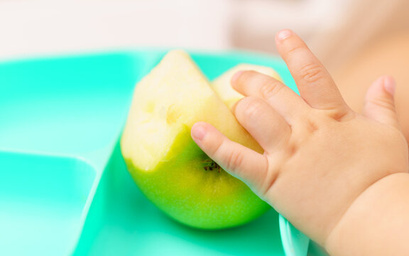 Close-up of a baby's hand exploring a partially eaten green apple on a child plate, representing feeding, independence and sensory development.