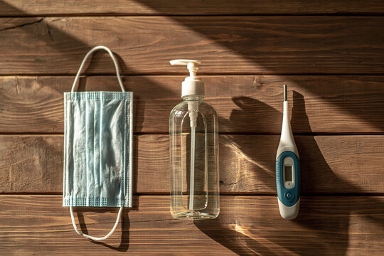 A medical face mask, sanitizer bottle, and digital thermometer sit on a rustic wooden table.