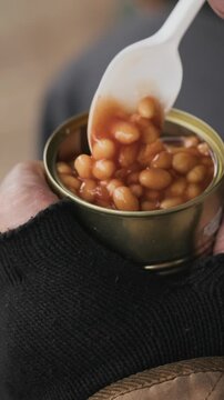 Vertical shot of homeless man eating canned beans with plastic spoon outdoors. Perfect for poverty awareness campaigns, social aid promos and charity projects