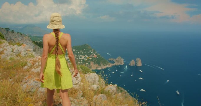 Woman walking on a high Capri cliff gazing over the coastline and iconic Faraglioni sea stacks below