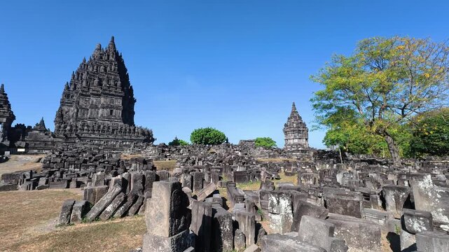 Weathered stone ruins fill the foreground beneath a Prambanan Temple tower and leafy tree in Central Java.