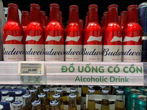 Red bottles of Budweiser arranged neatly on a store shelf. A sign indicating 'Alcoholic Drink' is displayed in the foreground. The scene suggests commercial retail and beverage marketing.