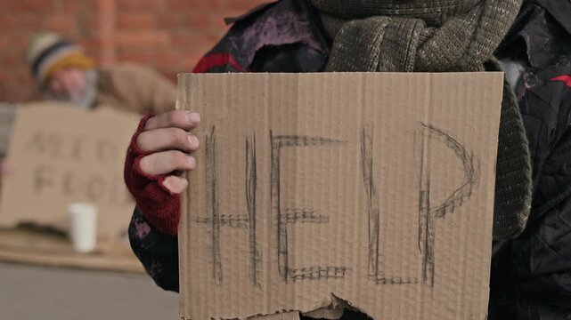 Portrait of young adult Caucasian homeless man holding cardboard help sign and begging tin can on street. Footage suitable for charity campaigns and social poverty awareness