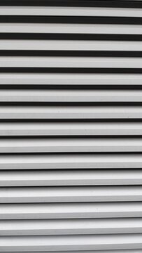 A modern industrial louvered vent in light gray, featuring a repeating geometric pattern of parallel horizontal lines and dark shadows.
