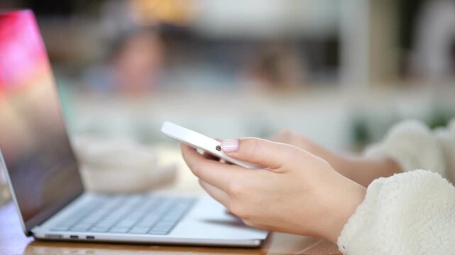Close-up of hands holding a smartphone. Laptop sits beside phone on wooden surface. Background blurred with warm indoor lighting. Person wears light-colored sweater. Focus stays on hands and devices
