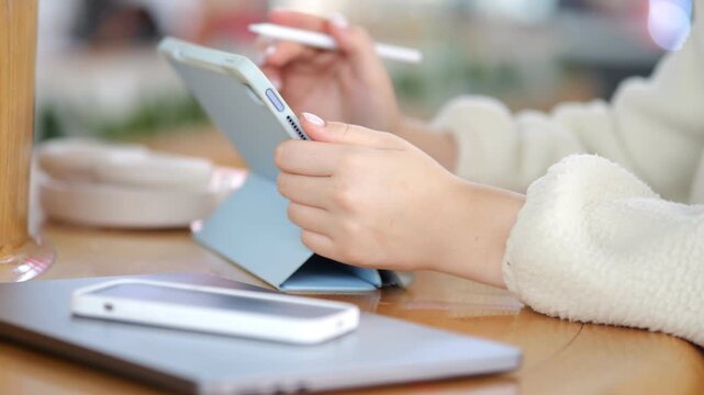Close-up of hands using tablet with stylus. Person wears soft white sweater. Table holds phone, laptop, and cup. Warm, cozy indoor setting with blurred background. Focus on digital creativity