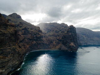 Mist drifts across the cliffs above an anchored yacht. Low mist catches on the upper slopes of Los Gigantes on western Tenerife. A small yacht rests on a patch of glittering water in the cove © Oksana