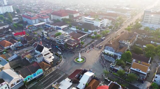 Aerial drone footage of Tugu Yogyakarta surrounded by a large urban intersection, city traffic, roads, and buildings during soft morning light in Yogyakarta, Indonesia.