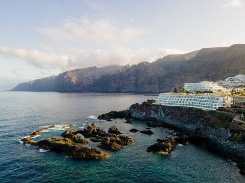 Basalt wall towers over a hotel and turquoise rock pools. The Los Gigantes cliff wall rises sheer from the Atlantic with a stepped white hotel on the headland. 