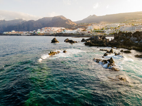 Aerial view over turquoise shallows toward a cliffside town. Drone view across clear turquoise water and scattered volcanic reefs toward the whitewashed town of Los Gigantes. The mountains of Tenerife