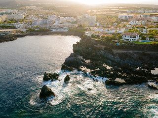 Rocky headland and resort villas above a glittering bay. Aerial view of a volcanic headland where waves wrap around submerged rocks below villas and apartment terraces. © Oksana