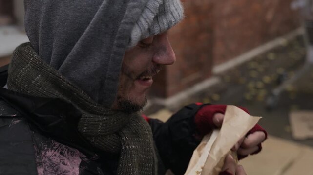 Caucasian homeless young adult male receiving food sandwich from volunteer and eating hungrily outdoors. Footage suits charity campaigns, food bank promotions, and poverty awareness projects
