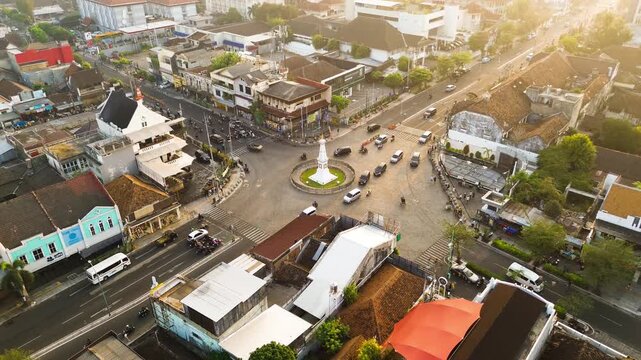 Aerial drone footage of Tugu Yogyakarta surrounded by urban roads, city buildings, traffic, and warm golden morning sunlight in Yogyakarta, Indonesia.
