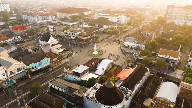 Aerial drone footage of Tugu Yogyakarta surrounded by urban roads, city buildings, traffic, and warm golden morning sunlight in Yogyakarta, Indonesia.