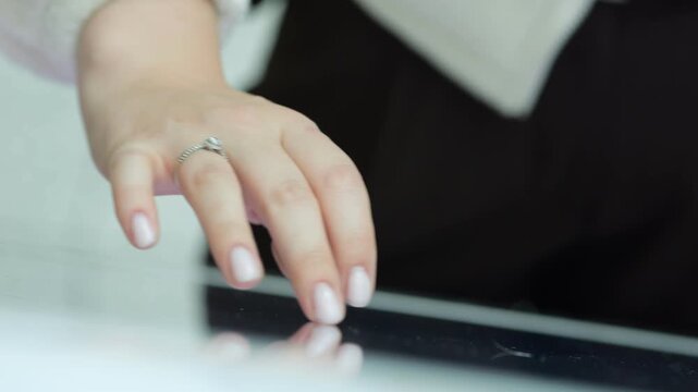 Close-up of a woman's hand touching a tablet screen. Her nails are neatly manicured with light polish. She wears a ring on her ring finger. The background is softly blurred with neutral tones