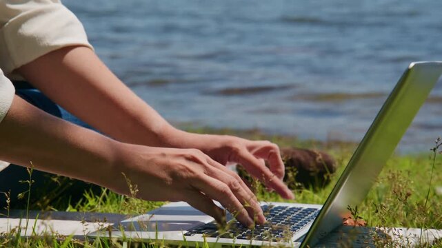 Person types on laptop near water. Hands show focused, calm work outdoors. Ideal for remote work, nature study, or digital lifestyle themes