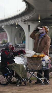 Vertical shot of Caucasian senior homeless man and man with disability in wheelchair drinking together under bridge. Suitable for charity and social support campaigns
