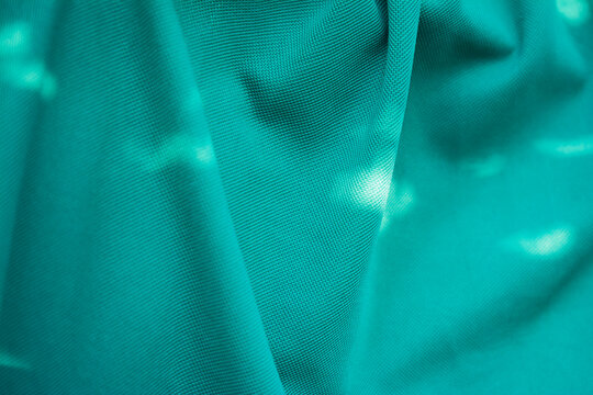 Close-up turquoise green fabric texture with flowing wavy folds, soft sunlight and tree shadows creating an abstract natural pattern.