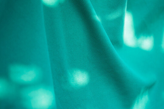Close-up turquoise green fabric texture with flowing wavy folds, soft sunlight and tree shadows creating an abstract natural pattern.