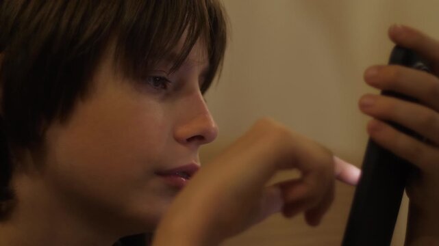 Close-up profile of a teenager's face intently looking at a smartphone screen in a dark room. The light from the gadget brightly illuminates the face of the engrossed child typing a message 