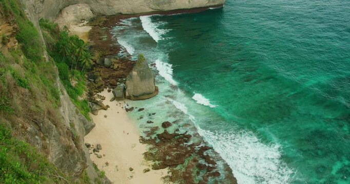 Palm trees grow along edge of towering cliffs at sandy shore Diamond beach in Nusa Penida Bali