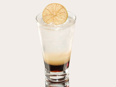 Tall glass of layered dark and clear soda drink with ice, topped with a dried lime wheel, shot on a light neutral background.