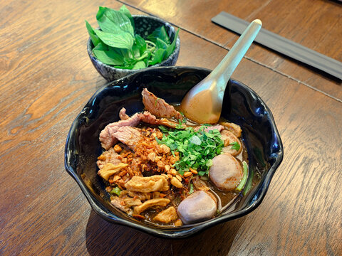 Close up of authentic Thai boat noodles with sliced meat, meatballs, and rich dark broth in a black bowl served with fresh basil on a wooden table