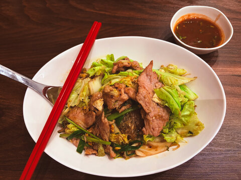 Top down view of Thai style dry sukiyaki stir-fried glass noodles with pork and cabbage in a white bowl served with spicy dipping sauce and red chopsticks