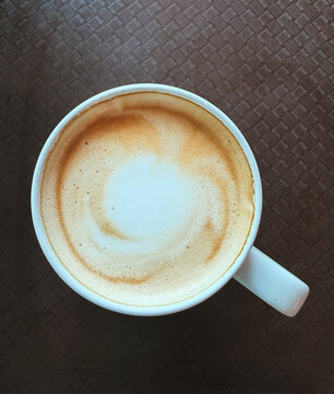 Top down close up view of a hot latte with frothy milk in a white ceramic mug on a brown textured table