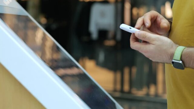 Man uses touchscreen kiosk while holding phone. Wears yellow shirt and green wristband. Background shows retail store interior. Hands interact with modern digital display