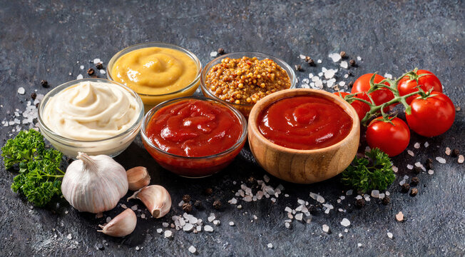 Different types of sauces in bowls with fresh ingredients on a dark surface